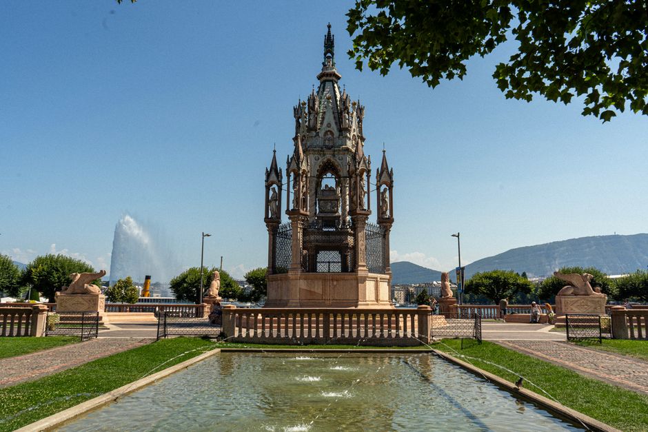 A historic ornate fountain with intricate carvings and decorative elements, situated in a landscaped park with a rectangular reflecting pool at its base. The fountain is surrounded by a paved walkway 
