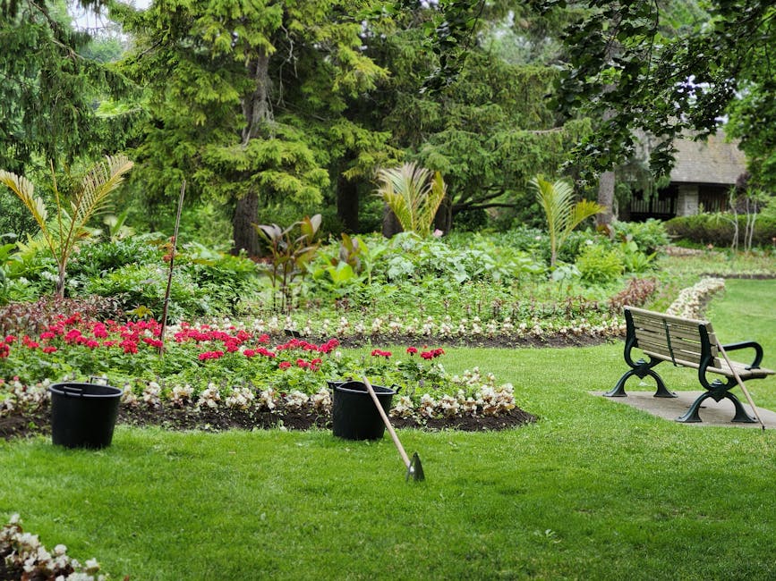 A well-maintained garden featuring vibrant flower beds with blooming pink and white flowers arranged in curved borders, surrounded by lush green grass. In the foreground, there are two black plastic b